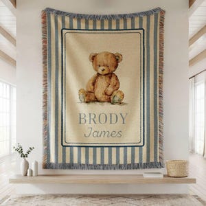 May include: A woven throw blanket featuring a teddy bear illustration. The blanket has a cream-colored background with blue vertical stripes and a rectangular frame around the bear. The name "Brody James" is printed below the bear.