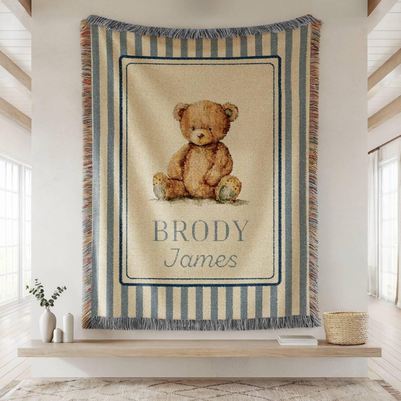 May include: A woven throw blanket featuring a teddy bear illustration. The blanket has a cream-colored background with blue vertical stripes and a rectangular frame around the bear. The name "Brody James" is printed below the bear.