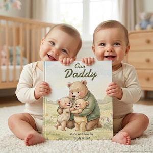 May include: Two smiling babies hold a book titled "Our Daddy." The book cover features a bear hugging two smaller bears, with text "Made with love by Teddy & Rae." The babies wear light-colored onesies and sit on a white rug.