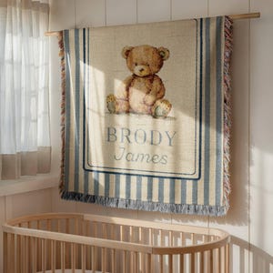 May include: A woven tapestry blanket featuring a brown teddy bear illustration. The blanket has blue and beige stripes and a blue border with the name "BRODY James" in the center. The blanket is hanging on a wooden rod.