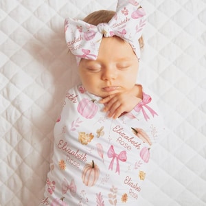 May include: A white swaddle blanket and matching headband set featuring a repeating pattern of pink pumpkins, bows, and autumn leaves. The fabric is printed with the name Elizabeth Rose in a cursive script throughout the design.