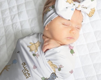 May include: A baby swaddled in a light gray blanket and wearing a matching headband, both adorned with Winnie the Pooh characters. The blanket is printed with Pooh, Piglet, Eeyore, and butterflies. The name "Olivia Claire" is printed on the blanket.
