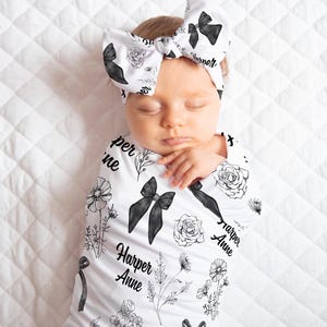 May include: A newborn baby wrapped in a white swaddle and wearing a matching bow headband. The swaddle and headband are decorated with black bows, floral designs, and the name "Harper Anne". The baby is lying on a white quilted surface.