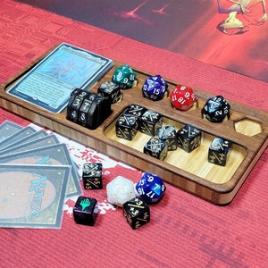 May include: A wooden dice tray with compartments for different sized dice. The tray is made of light brown wood with a dark brown border. There are several dice in the tray, including a black and white d20, a red d6, a blue d8, and a green d4. There is also a Magic: The Gathering card in the tray.