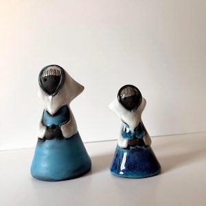 May include: Two ceramic figurines shaped like figures wearing hoods. The larger figurine has a light blue skirt and white hood, while the smaller one features a darker blue skirt and white hood. Both have dark brown faces with simple facial features.