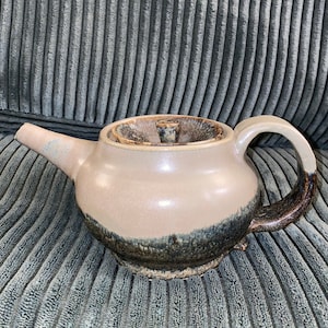 May include: A ceramic teapot with a brown and black glaze. The teapot has a round body and a lid with a knob. The teapot is sitting on a gray textured surface.