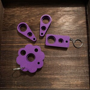 May include: Four purple keychains in various shapes: a rectangle, two teardrop shapes, and a flower. Each keychain has a circular hole and smaller holes. The rectangle keychain has a silver keyring attached. The items are on a dark wooden tray.