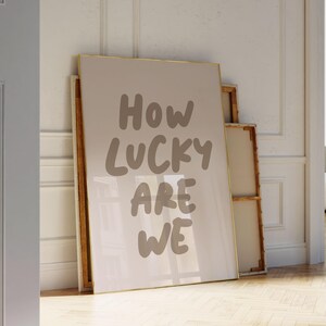 Puede incluir: Una impresi&oacute;n enmarcada con el texto "How lucky are we" en una fuente blanca manuscrita sobre un fondo beige.