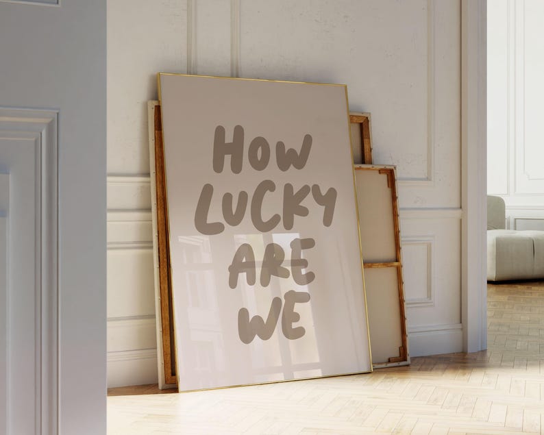 Puede incluir: Una impresi&oacute;n enmarcada con el texto "How lucky are we" en una fuente blanca manuscrita sobre un fondo beige.