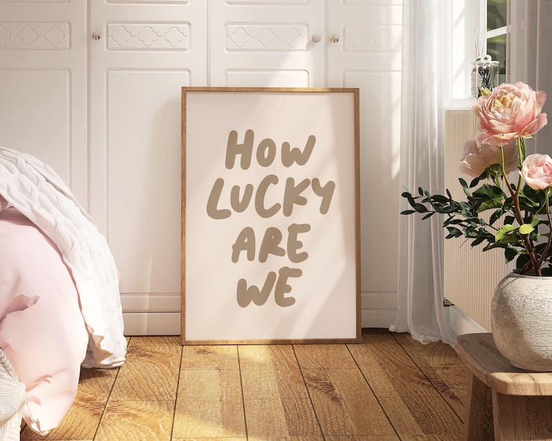 Puede incluir: Una impresi&oacute;n enmarcada con el texto "HOW LUCKY ARE WE" en una fuente manuscrita marr&oacute;n sobre un fondo rosa claro. La impresi&oacute;n est&aacute; colgada en una pared blanca en un dormitorio.