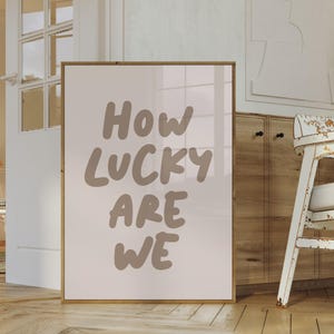 Puede incluir: Una impresi&oacute;n enmarcada con el texto "How lucky are we" en marr&oacute;n sobre un fondo blanco.