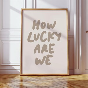Puede incluir: Una impresi&oacute;n enmarcada con un fondo rosa claro y el texto "How lucky are we" en una fuente de letra manuscrita marr&oacute;n.