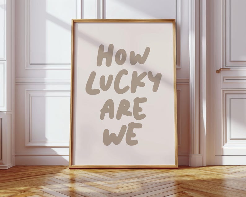 Puede incluir: Una impresi&oacute;n enmarcada con un fondo rosa claro y el texto "How lucky are we" en una fuente de letra manuscrita marr&oacute;n.