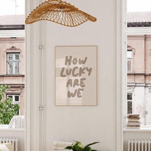 Puede incluir: Una impresi&oacute;n enmarcada beige con el texto "HOW LUCKY ARE WE" en una fuente de pincel marr&oacute;n. La impresi&oacute;n est&aacute; colgada en una pared blanca en una habitaci&oacute;n con dos ventanas y una l&aacute;mpara de tejido.