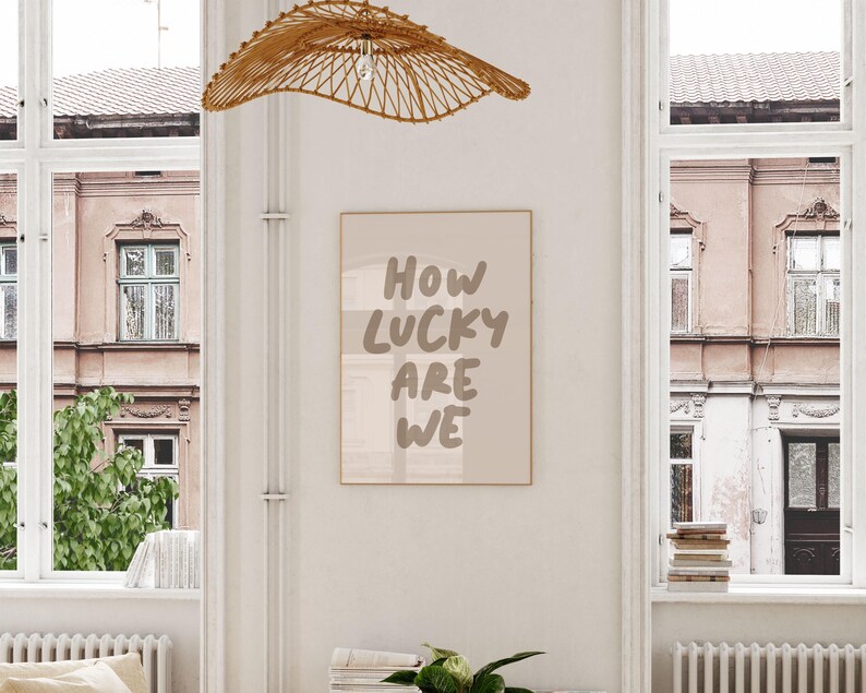Puede incluir: Una impresi&oacute;n enmarcada beige con el texto "HOW LUCKY ARE WE" en una fuente de pincel marr&oacute;n. La impresi&oacute;n est&aacute; colgada en una pared blanca en una habitaci&oacute;n con dos ventanas y una l&aacute;mpara de tejido.