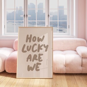 Puede incluir: Una impresi&oacute;n enmarcada con el texto "HOW LUCKY ARE WE" en una fuente manuscrita marr&oacute;n. La impresi&oacute;n est&aacute; colgada en una pared en una habitaci&oacute;n con un sof&aacute; rosa y una ventana con vista a un horizonte urbano.