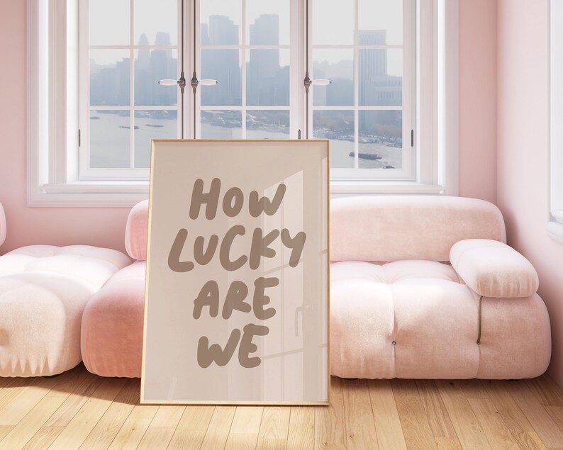 Puede incluir: Una impresi&oacute;n enmarcada con el texto "HOW LUCKY ARE WE" en una fuente manuscrita marr&oacute;n. La impresi&oacute;n est&aacute; colgada en una pared en una habitaci&oacute;n con un sof&aacute; rosa y una ventana con vista a un horizonte urbano.