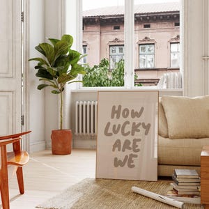 Puede incluir: Un p&oacute;ster enmarcado en beige con el texto "HOW LUCKY ARE WE" en una fuente manuscrita marr&oacute;n. El p&oacute;ster est&aacute; apoyado en un sof&aacute; beige con un coj&iacute;n. Una planta de ficus en maceta est&aacute; en el fondo.