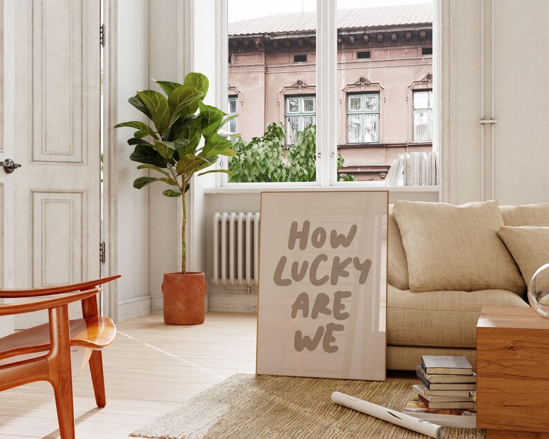 Puede incluir: Un p&oacute;ster enmarcado en beige con el texto "HOW LUCKY ARE WE" en una fuente manuscrita marr&oacute;n. El p&oacute;ster est&aacute; apoyado en un sof&aacute; beige con un coj&iacute;n. Una planta de ficus en maceta est&aacute; en el fondo.