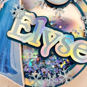 Puede incluir: Adorno para tarta circular en capas con el nombre "Elyse" en letras plateadas iridiscentes. El adorno presenta una combinaci&oacute;n de colores azul y plateado, con detalles de copos de nieve y un centro transparente lleno de purpurina. Imagen de dibujos animados.