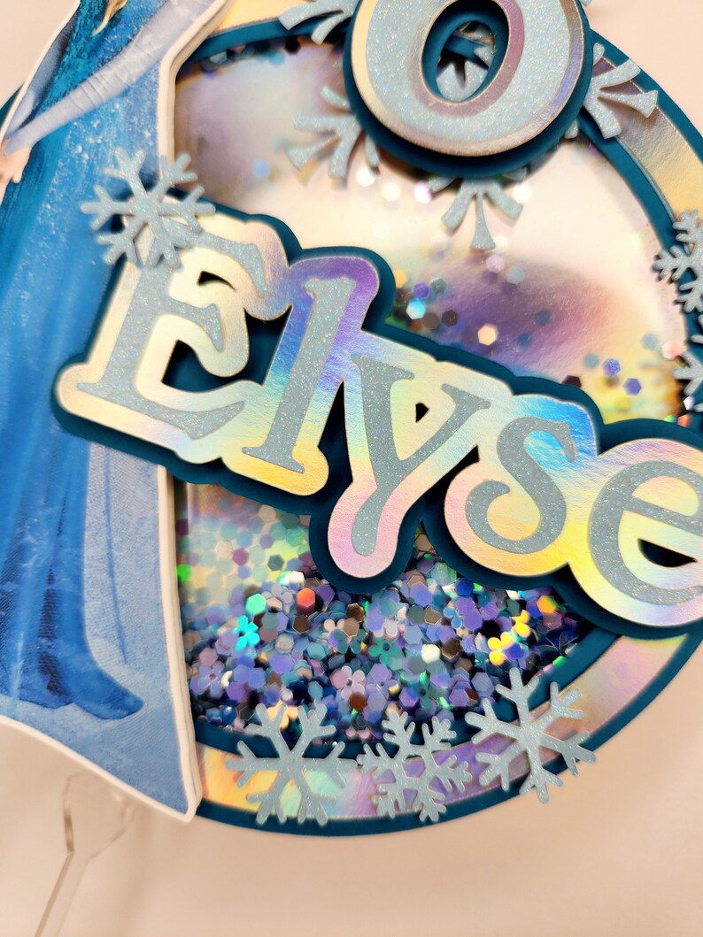 Puede incluir: Adorno para tarta circular en capas con el nombre "Elyse" en letras plateadas iridiscentes. El adorno presenta una combinaci&oacute;n de colores azul y plateado, con detalles de copos de nieve y un centro transparente lleno de purpurina. Imagen de dibujos animados.