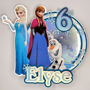 Puede incluir: Adorno para tarta con Elsa, Anna y Olaf de Frozen. El dise&ntilde;o incluye un fondo azul y plateado con purpurina, el n&uacute;mero 6, copos de nieve y el nombre "Elyse". Los personajes visten sus trajes caracter&iacute;sticos.