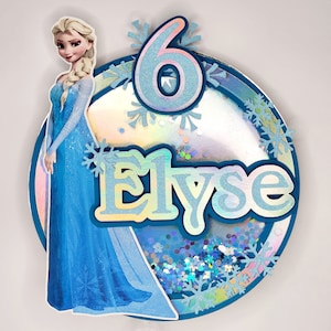 Puede incluir: Un adorno para tarta personalizado con el personaje de Elsa de Frozen. El adorno redondo tiene un borde azul, el nombre "Elyse", el n&uacute;mero "6" y detalles de copos de nieve. El fondo est&aacute; lleno de purpurina iridiscente.
