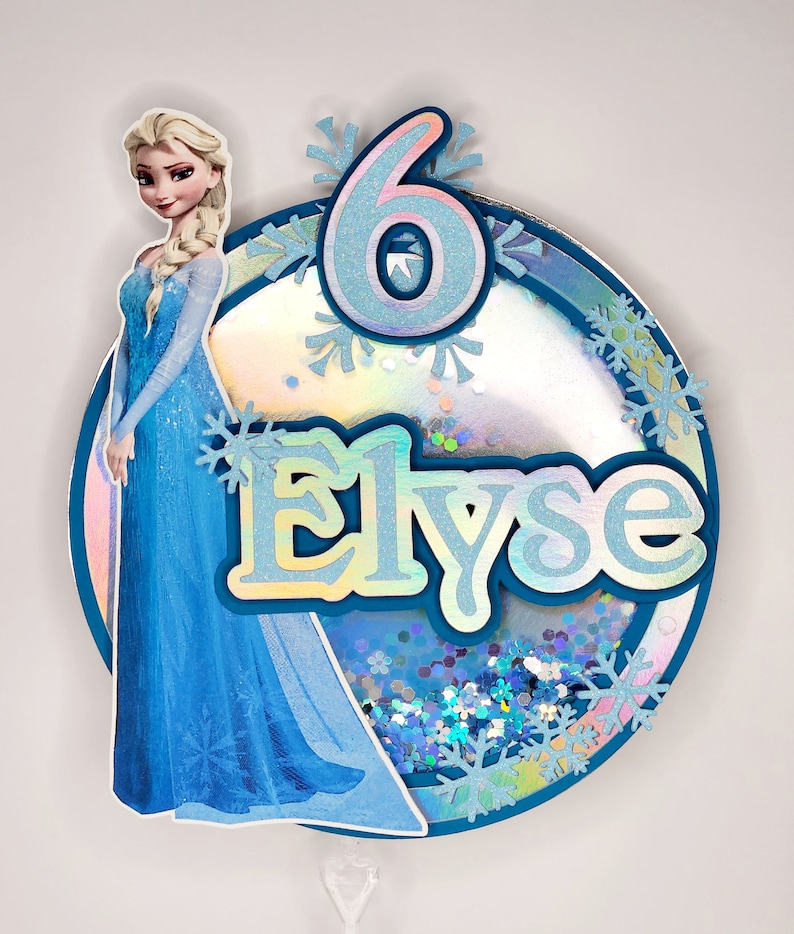 Puede incluir: Un adorno para tarta personalizado con el personaje de Elsa de Frozen. El adorno redondo tiene un borde azul, el nombre "Elyse", el n&uacute;mero "6" y detalles de copos de nieve. El fondo est&aacute; lleno de purpurina iridiscente.