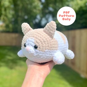 May include: A crocheted cat plushie in cream, tan, and white. The cat has black eyes and a pink nose. The plushie is round and has small ears and paws. The image includes a circular graphic that says "PDF Pattern Only".