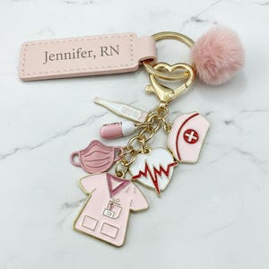Pode incluir: Um porta-chaves dourado com uma etiqueta retangular de couro rosa claro gravada com o texto Jennifer, RN. Inclui um pompom rosa macio e vários berloques de esmalte com tema médico, incluindo uma blusa de uniforme, máscara, ritmo cardíaco, pílula e touca.