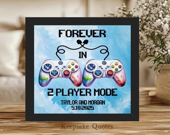 May include: Framed artwork with a black border featuring two colorful game controllers connected by a cable. Text reads "FOREVER IN 2 PLAYER MODE" with names and a date below. The background is a light blue watercolor design.