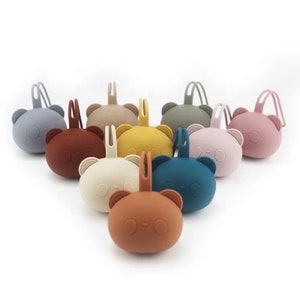 Könnte beinhalten: Set mit zehn Silikon-Beißringen in Form von Bären in verschiedenen Farben, darunter Braun, Blau, Gelb, Grau und Rosa. Jeder Beißring hat eine Schlaufe zum Befestigen an einem Kinderwagen oder Autositz.