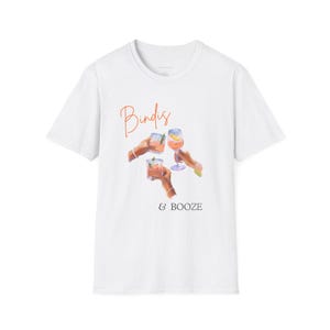 May include: White t-shirt with the words "Bindis & BOOZE" in orange script. The shirt features a watercolor illustration of three hands holding cocktails. The drinks are in clear glasses and have orange and yellow hues.