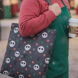 May include: A tote bag with a Jack Skellington pattern, featuring a black background with white Jack faces, Christmas trees, and snowflakes. The bag is held by a person wearing a red shirt and green apron, holding a coffee cup.
