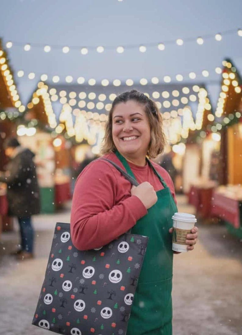 May include: A tote bag with a Jack Skellington pattern, featuring a black background with white Jack faces, Christmas trees, and snowflakes. The bag is held by a person wearing a red shirt and green apron, holding a coffee cup.