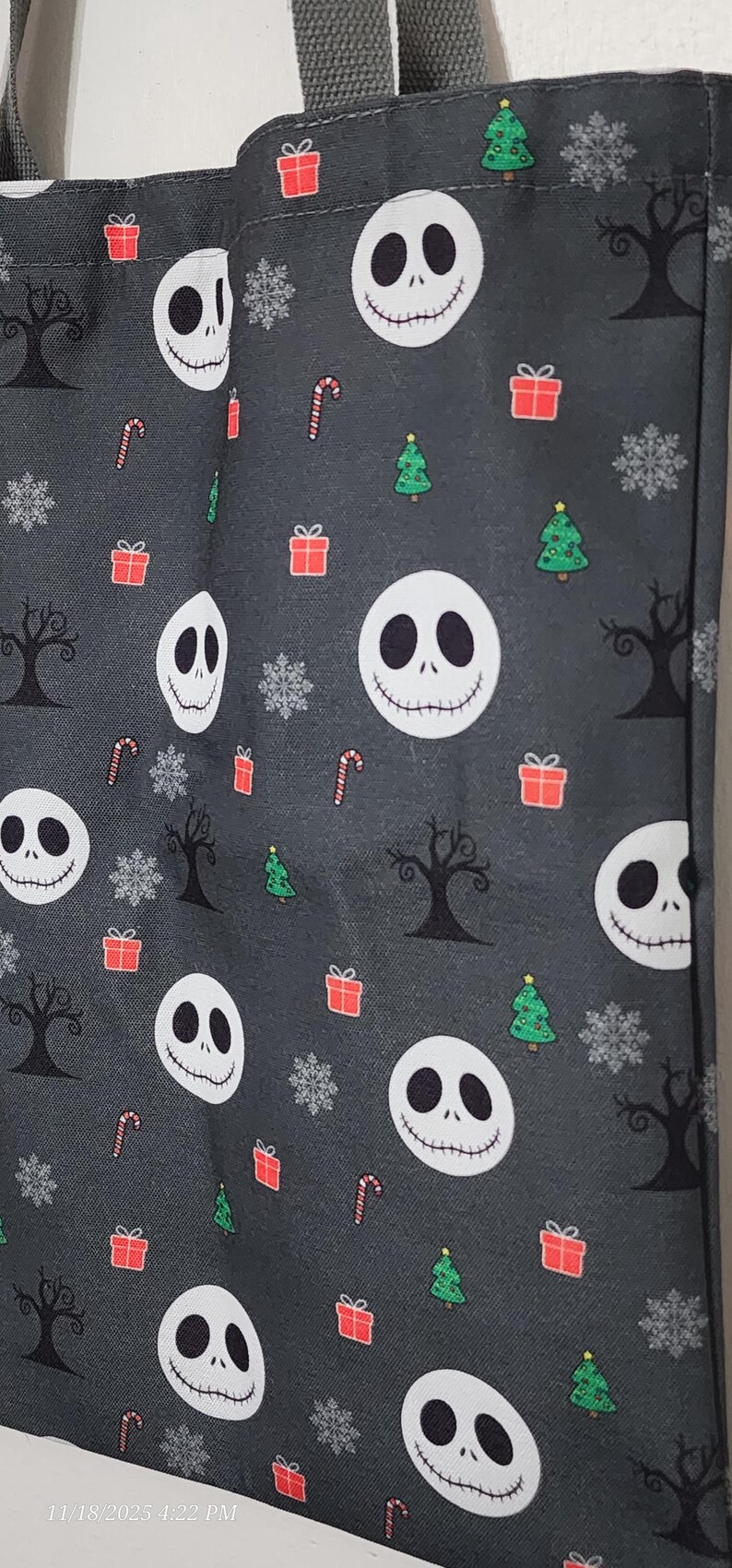 May include: A dark gray tote bag featuring a repeating pattern of Jack Skellington faces, Christmas trees, snowflakes, candy canes, and wrapped gifts. The bag has a gray handle and is made of a durable fabric.