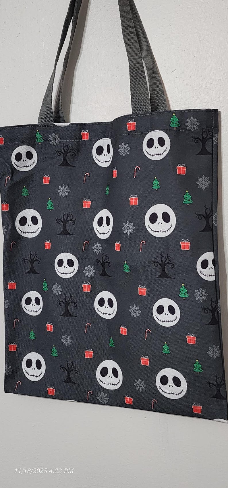 May include: A gray tote bag featuring a pattern of Jack Skellington faces, Christmas trees, snowflakes, gift boxes, and bare trees. The bag has gray handles and is likely made of fabric.