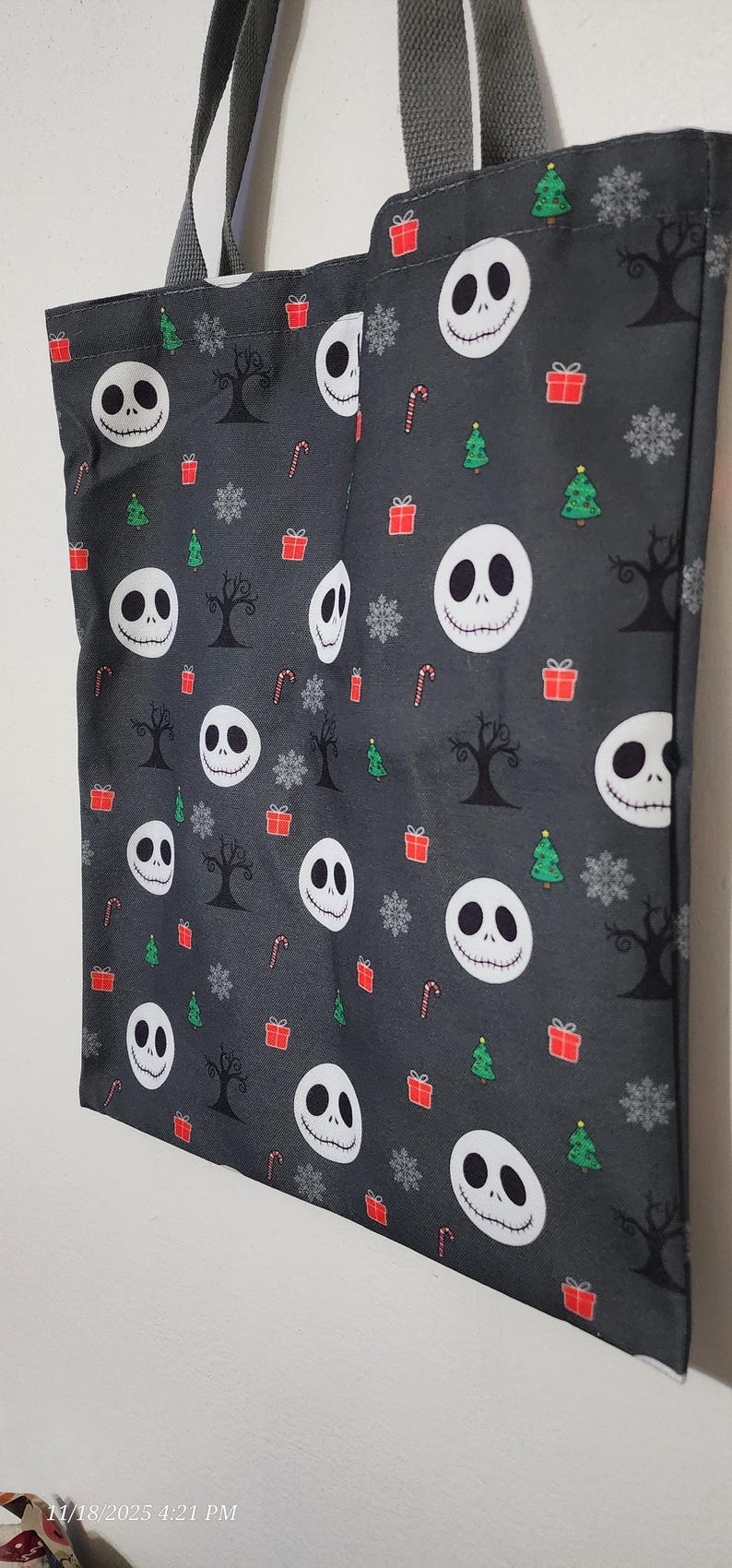 May include: A gray tote bag featuring a pattern of Jack Skellington faces, Christmas trees, snowflakes, candy canes, and gift boxes. The bag has gray handles and is made of fabric.