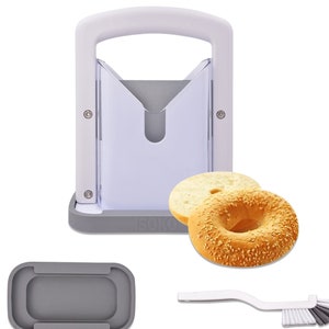 May include: White plastic bagel slicer with a gray base and a gray silicone base. The slicer has a handle and a cutting mechanism. Two bagels are shown next to the slicer. One bagel is plain and the other has sesame seeds. A gray silicone cleaning brush and a gray silicone base are also shown.