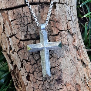 May include: A silver chain necklace with a silver cross pendant. The cross has a geometric design with sharp angles. The necklace is lying on a piece of weathered wood.