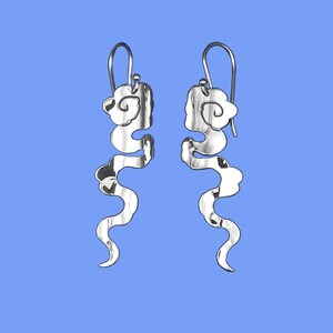 May include: A pair of silver earrings with a cloud-like design. The earrings have a hook closure and a flowing, abstract shape. The design features interconnected cloud shapes and a wavy, elongated form. The earrings are displayed against a blue background.