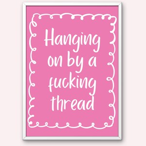 Puede incluir: Impresión digital rosa enmarcada con la frase en blanco « Hanging on by a fucking thread ». La impresión tiene un borde blanco y está sobre un fondo rosa claro. Las palabras « DIGITAL PRINT » están en la parte inferior.