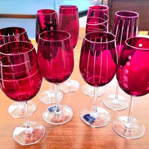 May include: A collection of ruby red wine glasses with various etched designs, including stripes and polka dots. The glasses have clear stems and bases, arranged on a light brown surface. The glasses are for serving wine.