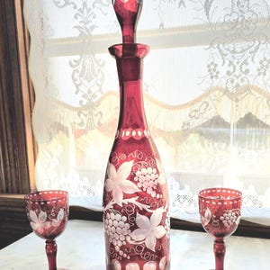 Puede incluir: Una jarra de cristal rojo rubí con tapón a juego y dos copas de vino pequeñas. La jarra y las copas presentan un diseño de uvas y hojas blancas. La jarra mide aproximadamente 30 cm de altura.