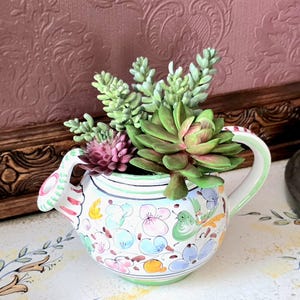 May include: A hand-painted ceramic teapot planter filled with faux succulents. The teapot is white with colorful floral and bird designs, a green handle, and a green rim. The succulents are various shades of green and purple.