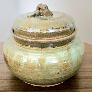 May include: A handmade ceramic jar with a lid. The jar is a muted green with brown and tan accents. The lid has a small, textured brown knob. The jar is round and has a rustic, organic feel.