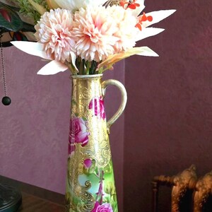 May include: A tall, decorative vase with a handle, filled with artificial flowers. The vase is gold with a floral design featuring pink roses and green leaves. The flowers are a mix of white, peach, and red.