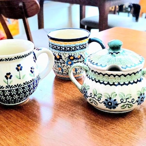 May include: Three ceramic pieces with floral patterns sit on a wooden table. The set includes two mugs and a small sugar bowl with a lid. The mugs and sugar bowl have blue, green, and white designs.