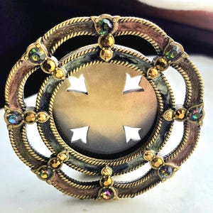 May include: A circular brooch featuring an antique brass-toned metal frame with a rope-textured border. The center contains a cutout cross shape against a dark gradient background. The frame is accented with small iridescent and amber-colored crystals.
