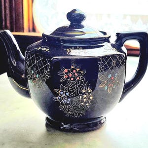 May include: A dark blue ceramic teapot with a lid and handle. The teapot is decorated with floral and geometric patterns in gold, red, and white. The spout and handle have gold trim. A decorative item for tea lovers.